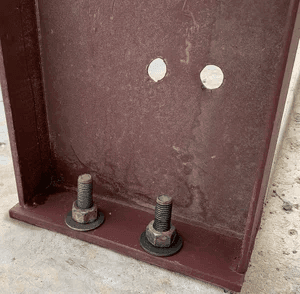 Red iron steel building base plate with anchor bolts and welded I-beam column secured to concrete foundation.