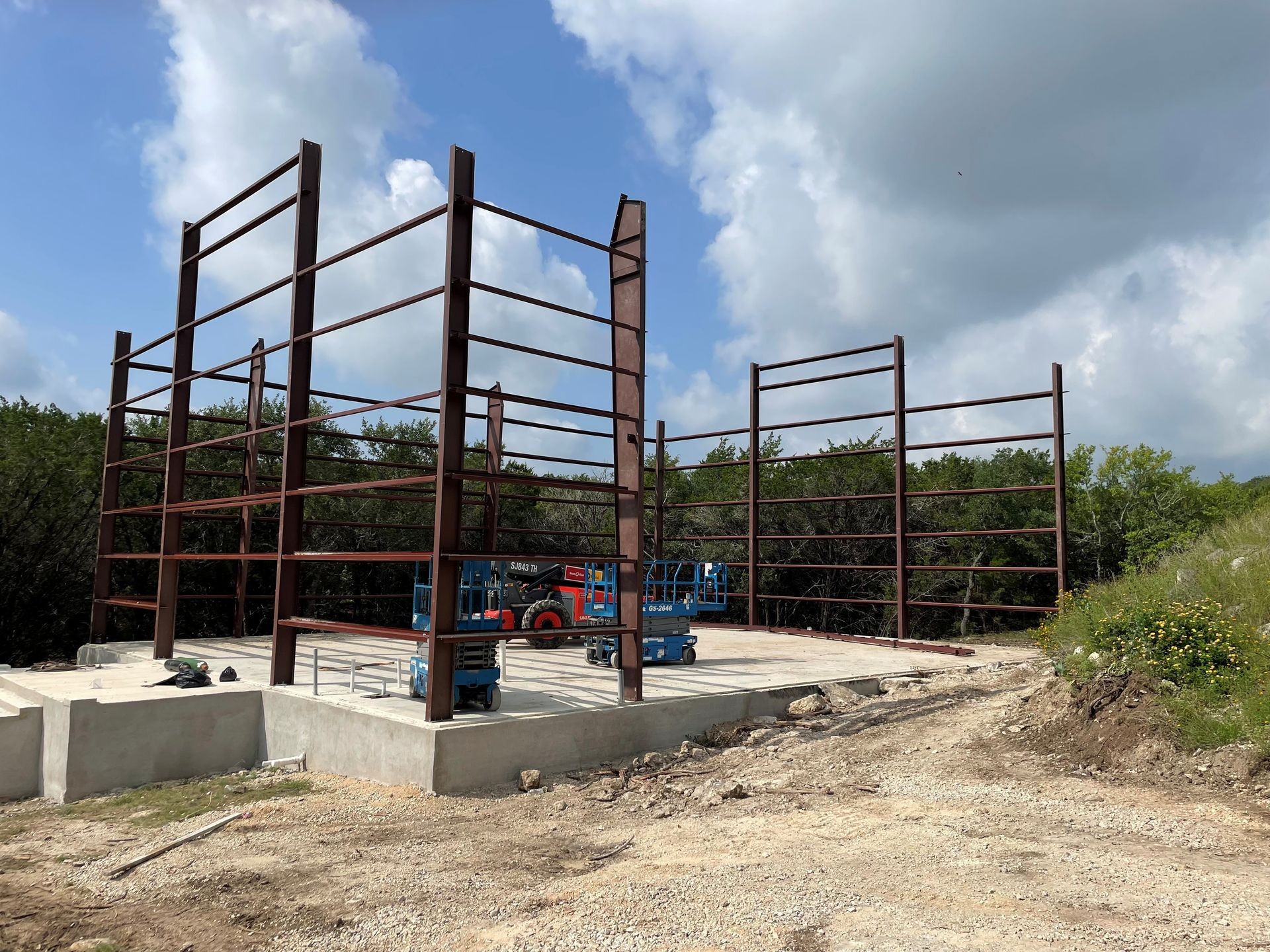 Red iron metal building frame under construction on concrete slab in rural Texas – pre-engineered steel structure installation by Texas Metal Buildings LLC